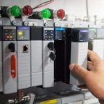 Hand adjusting a PLC module inside an industrial control panel with indicator lights.