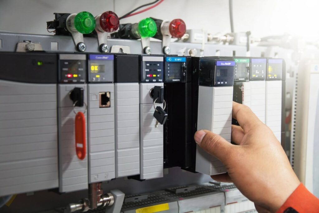 Hand adjusting a PLC module inside an industrial control panel with indicator lights.