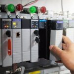 Hand adjusting a PLC module inside an industrial control panel with indicator lights.