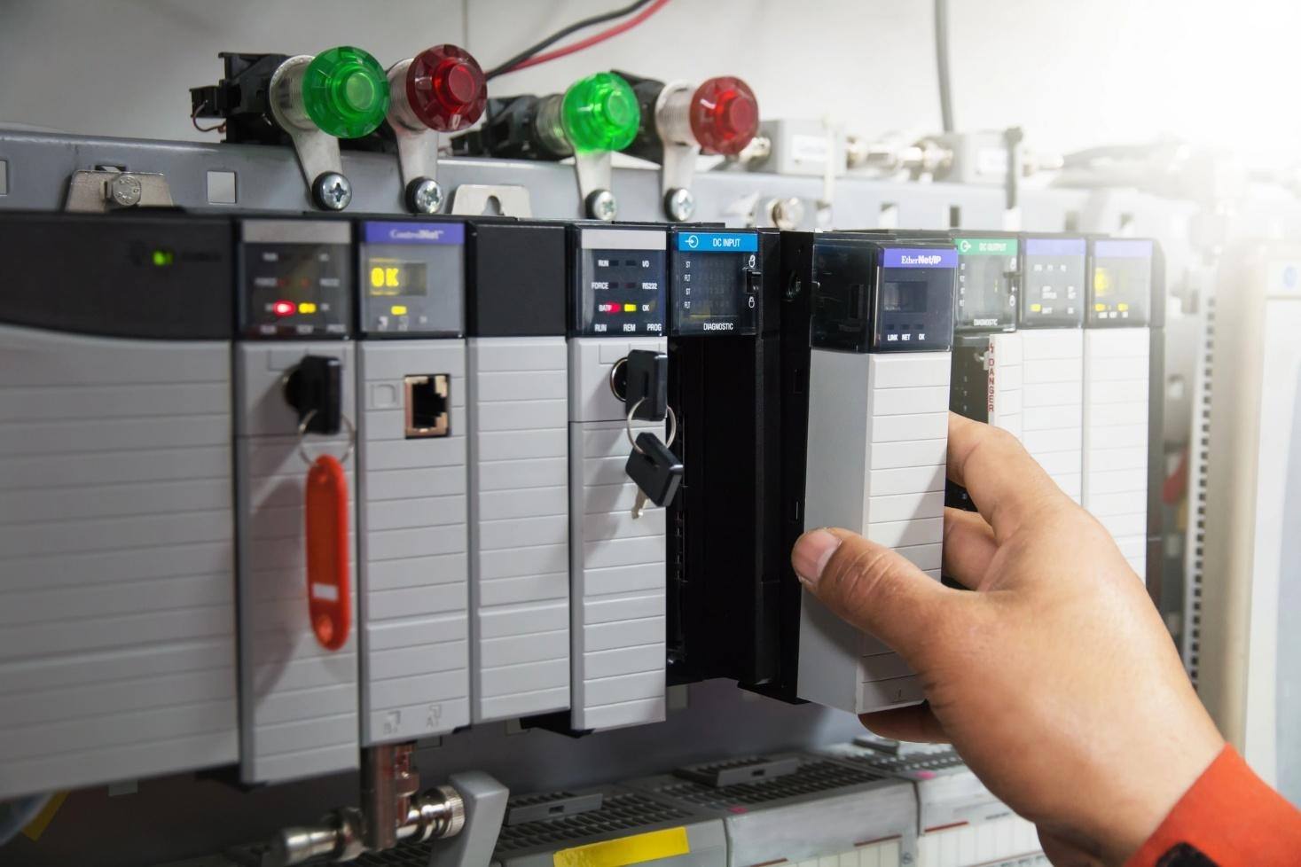 Hand adjusting a PLC module inside an industrial control panel with indicator lights.