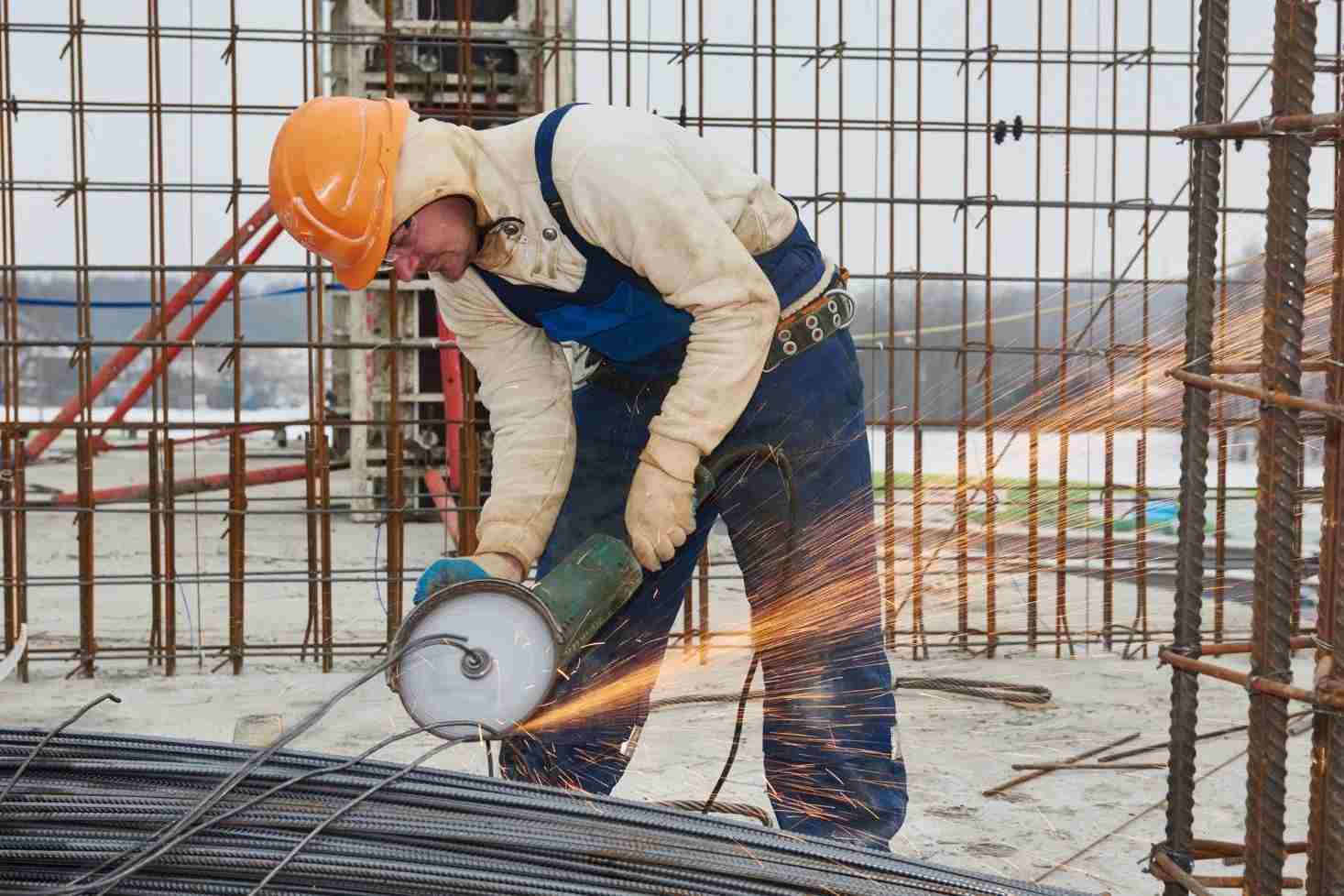 Worker using a handheld grinder to cut steel rebar, producing sparks at an active construction site.